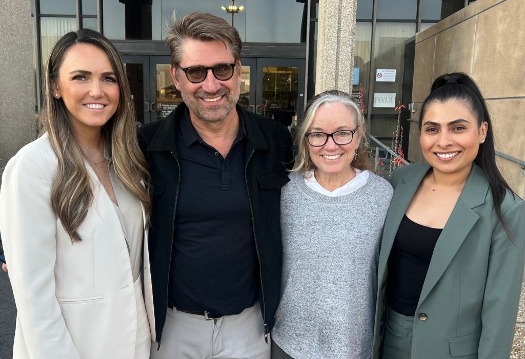 Four adults, three women and one man, stand close together and smile for the camera outside a building with glass doors and concrete walls.