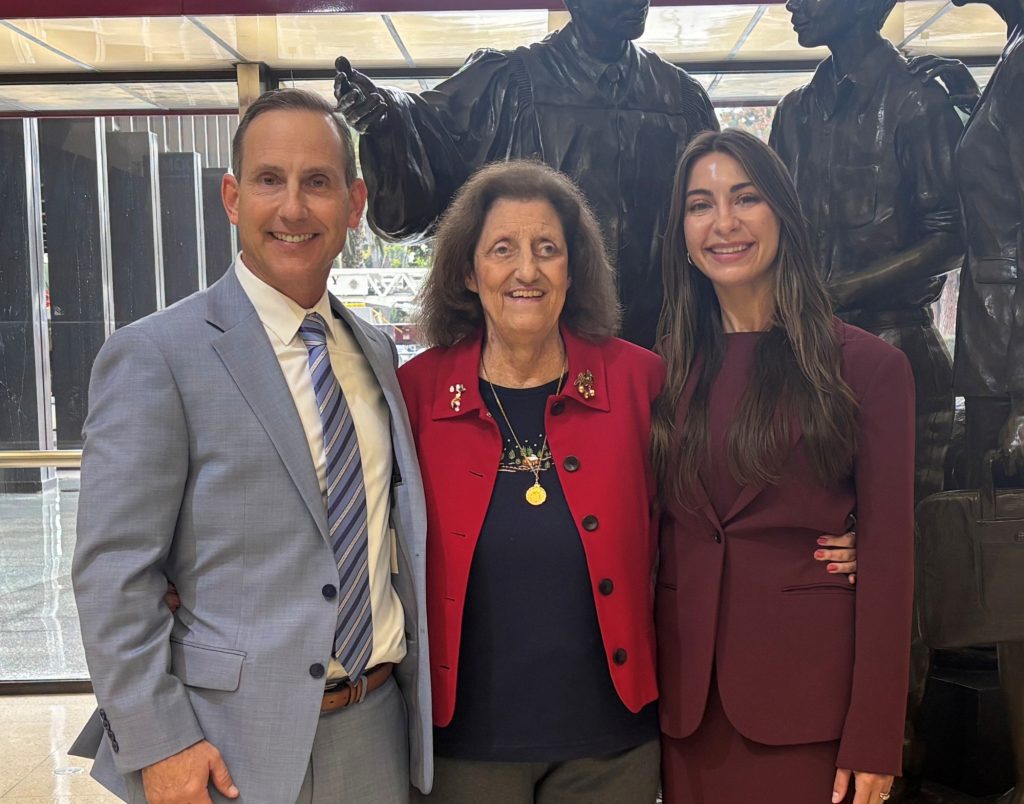Three people stand close together and smile for the camera; a man in a light suit, an older woman in a red jacket, and a woman in a dark suit, with statues and large windows in the background.