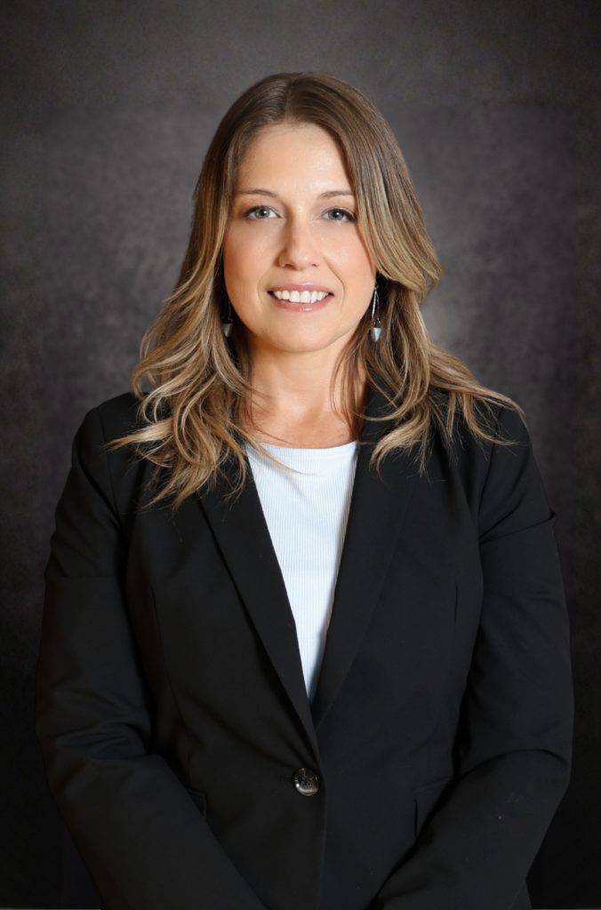 A woman with long, blonde hair wearing a black blazer and white top stands in front of a dark, textured background and smiles at the camera.
