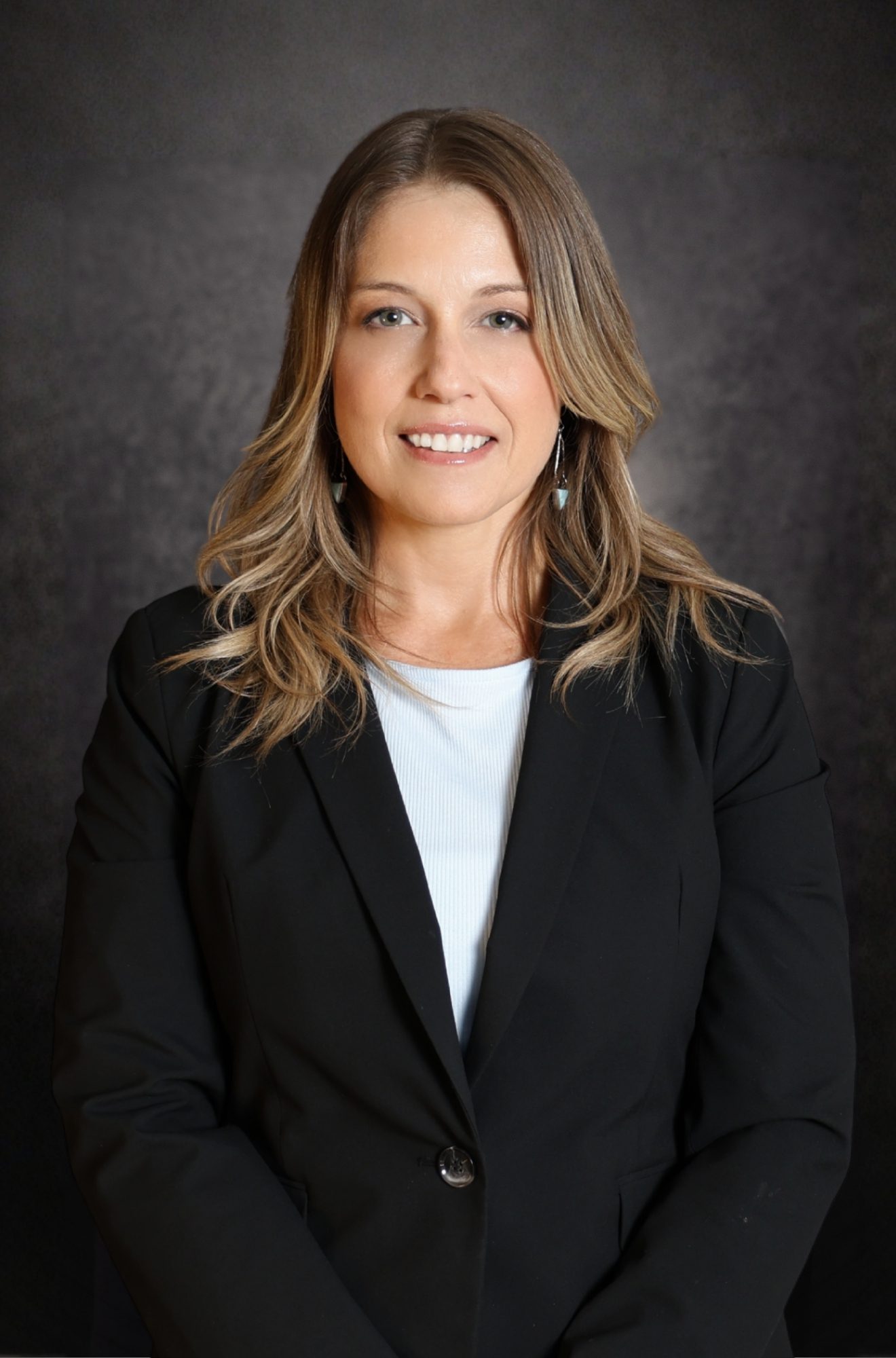 A woman with long, blonde hair wearing a black blazer and white top stands in front of a dark, textured background and smiles at the camera.
