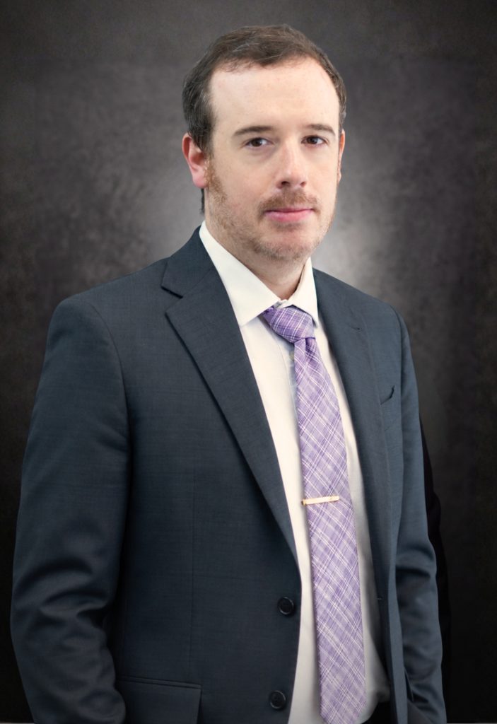A man wearing a dark suit, white shirt, and light purple patterned tie stands in front of a dark, textured background, looking at the camera with a neutral expression.