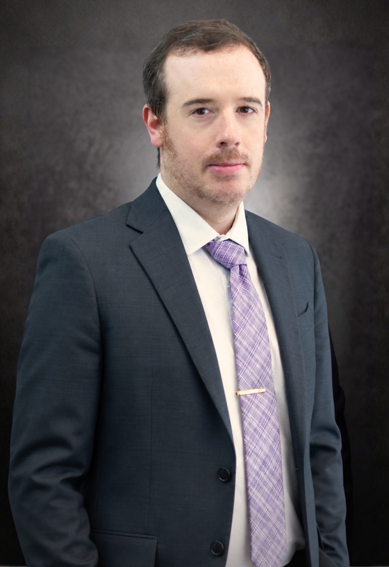 A man wearing a dark suit, white shirt, and light purple patterned tie stands in front of a dark, textured background, looking at the camera with a neutral expression.