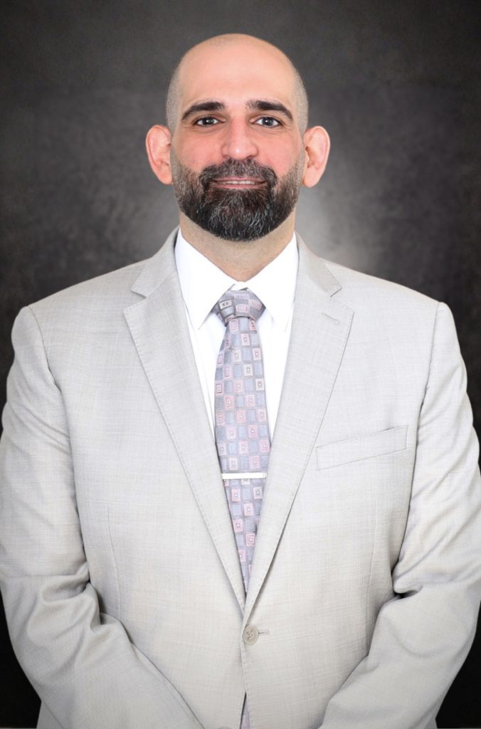 A man with a bald head and beard is wearing a light gray suit, patterned tie, and white shirt, standing against a dark background, facing the camera and smiling slightly.