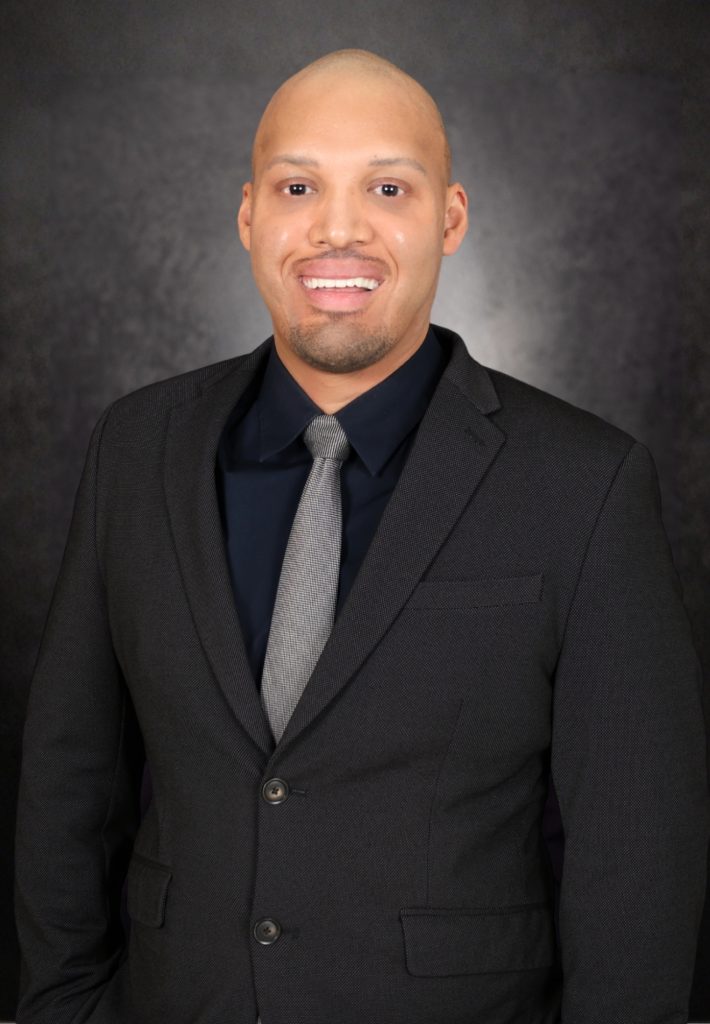 A man in a dark suit and gray tie stands smiling against a dark, textured background. He has a shaved head and no facial hair.