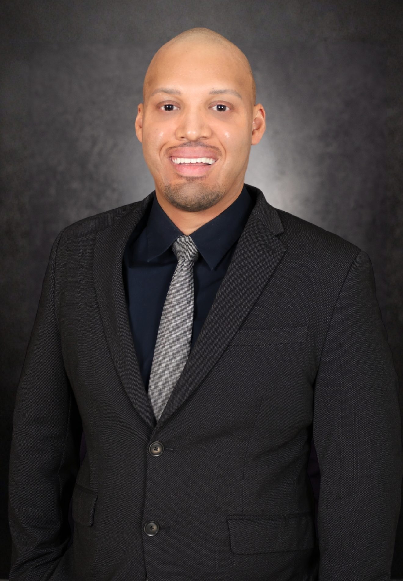 A man in a dark suit and gray tie stands smiling against a dark, textured background. He has a shaved head and no facial hair.