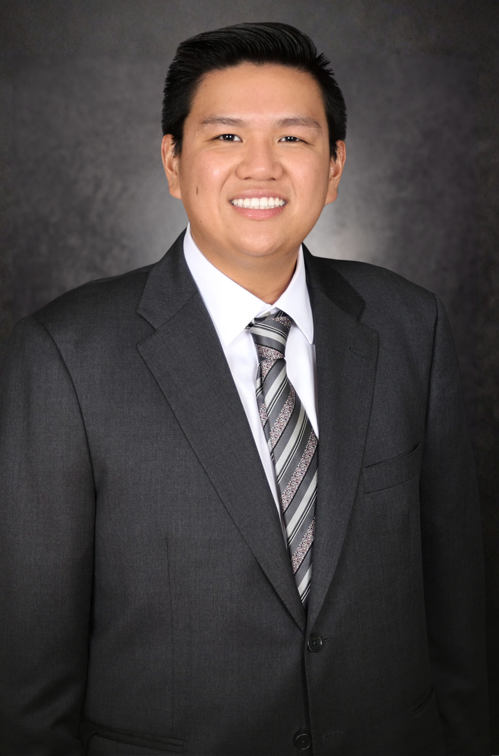 A man wearing a dark suit, white shirt, and striped tie stands smiling in front of a dark, textured background.