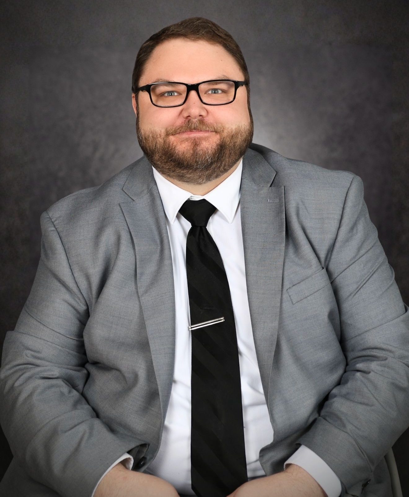A man with glasses, a beard, and short hair is wearing a gray suit, white shirt, and black tie. He is sitting against a dark background and facing the camera with a neutral expression.