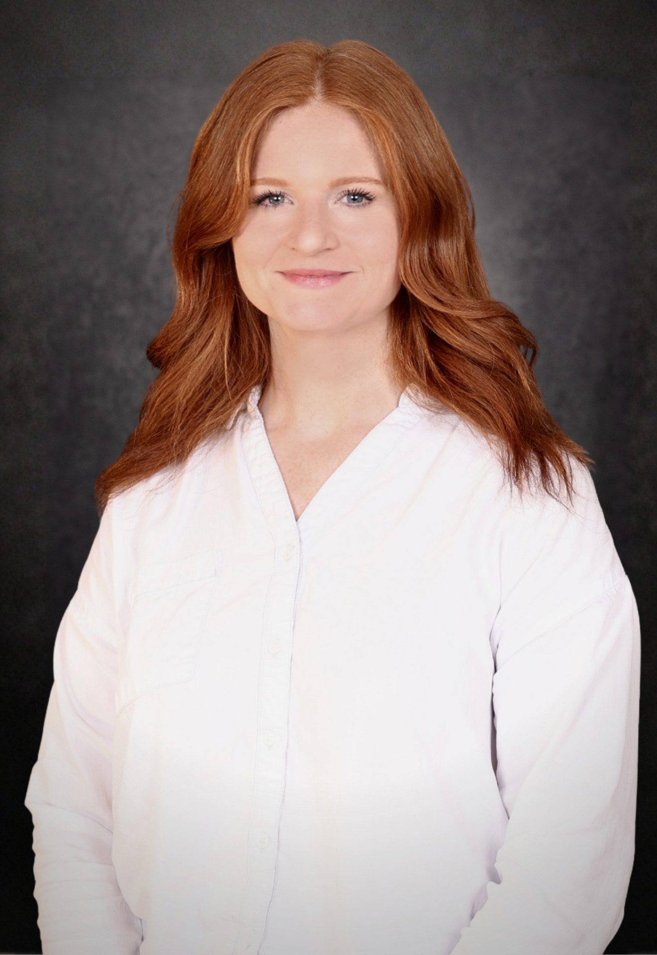A woman with long red hair, wearing a white button-up shirt, stands in front of a dark, textured background and smiles at the camera.