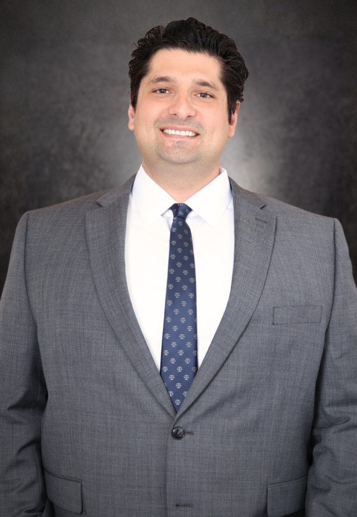 A man in a gray suit, white shirt, and patterned tie is smiling at the camera. He has dark, wavy hair and stands against a dark, textured background.