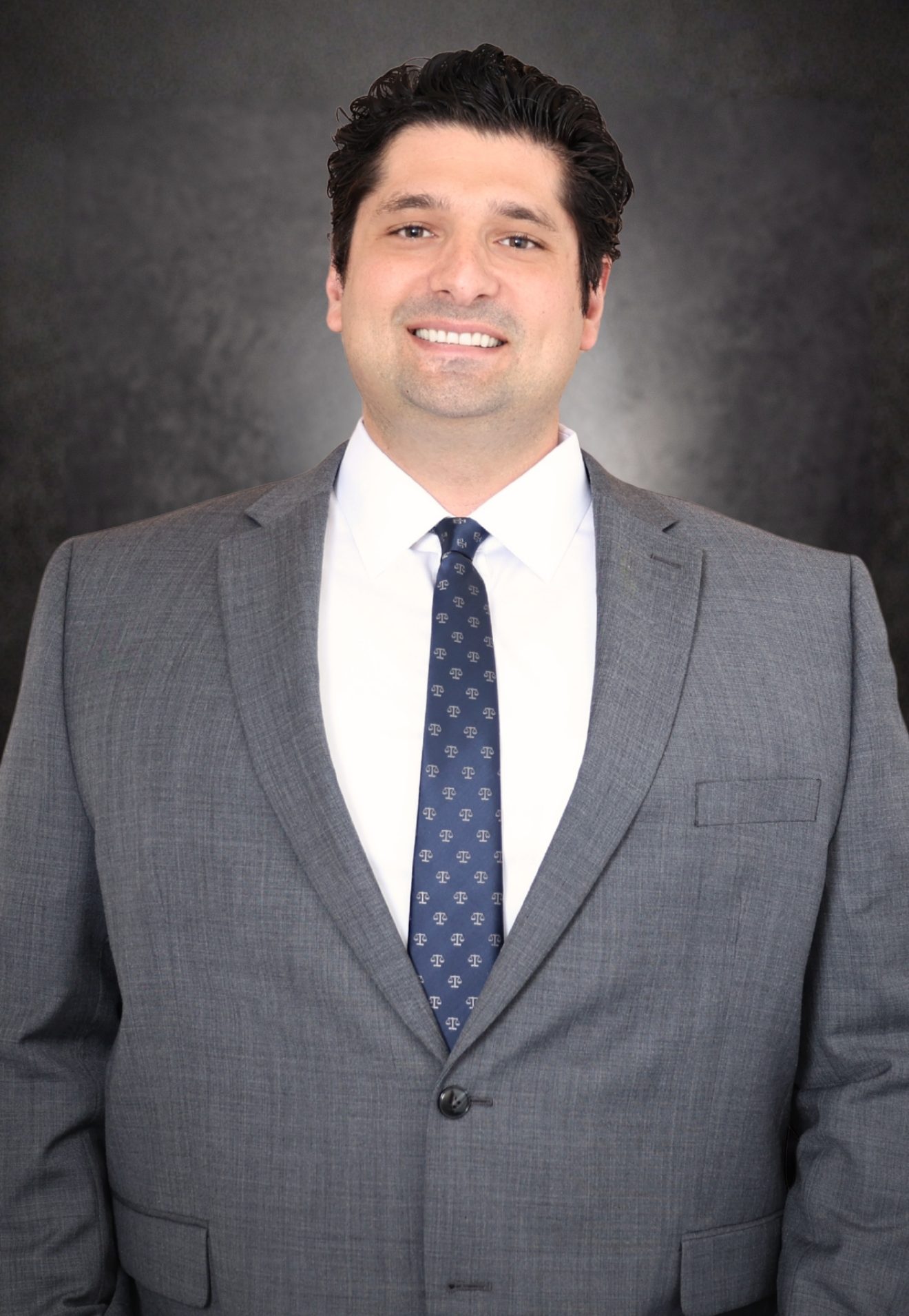 A man in a gray suit, white shirt, and patterned tie is smiling at the camera. He has dark, wavy hair and stands against a dark, textured background.