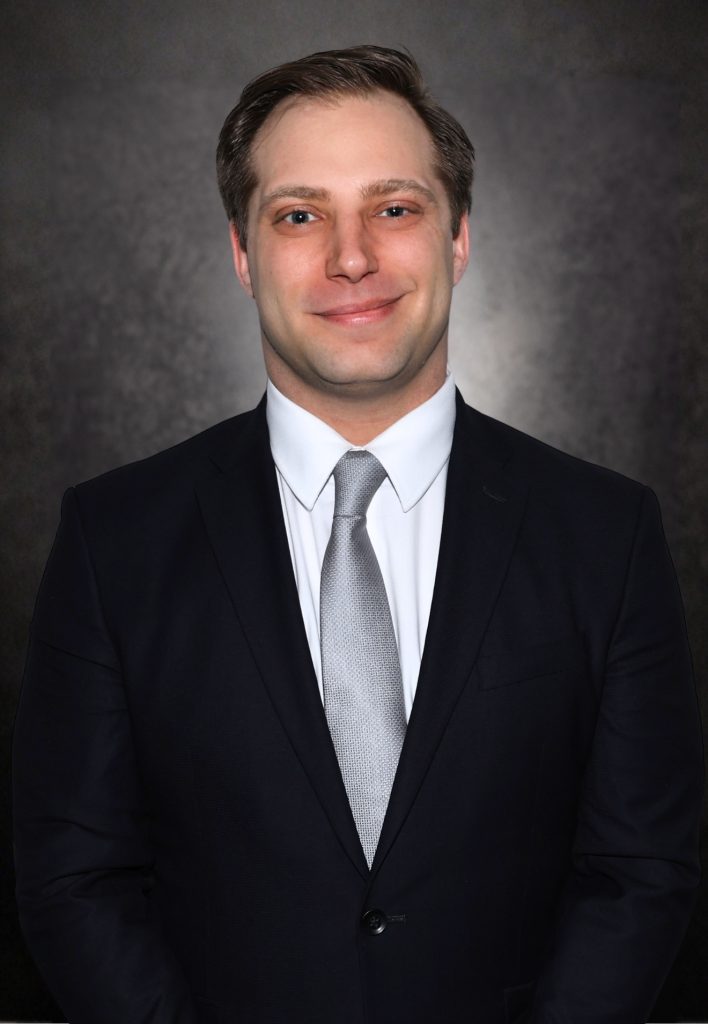 A man in a dark suit, white shirt, and silver tie stands against a dark, textured background, facing the camera and smiling slightly.