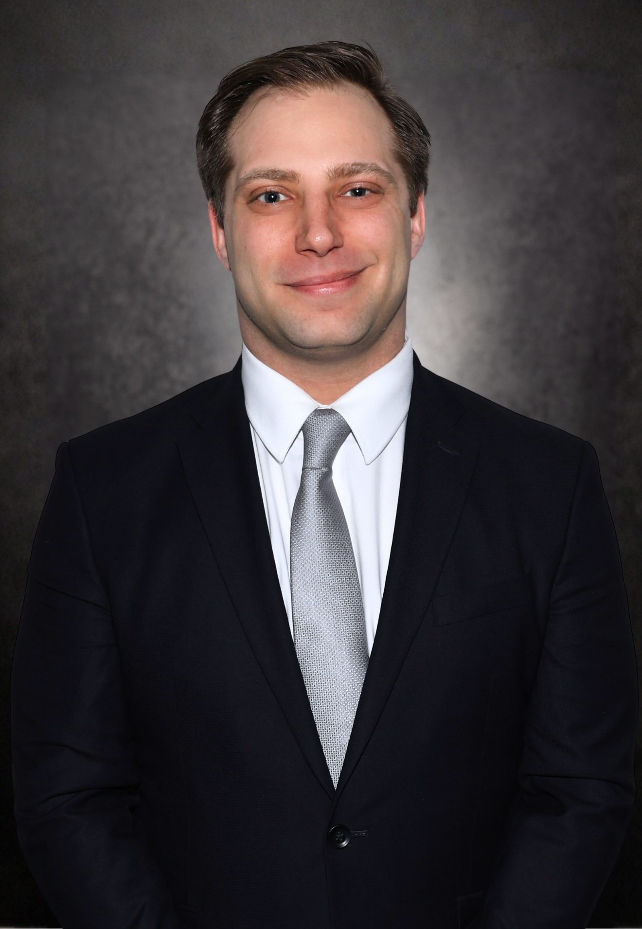 A man in a dark suit, white shirt, and silver tie stands against a dark, textured background, facing the camera and smiling slightly.