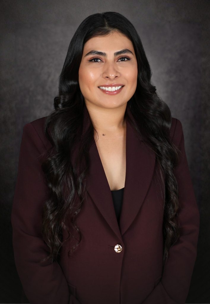 Woman with long dark hair wearing a burgundy blazer and a black top, smiling at the camera, with a dark, blurred background.
