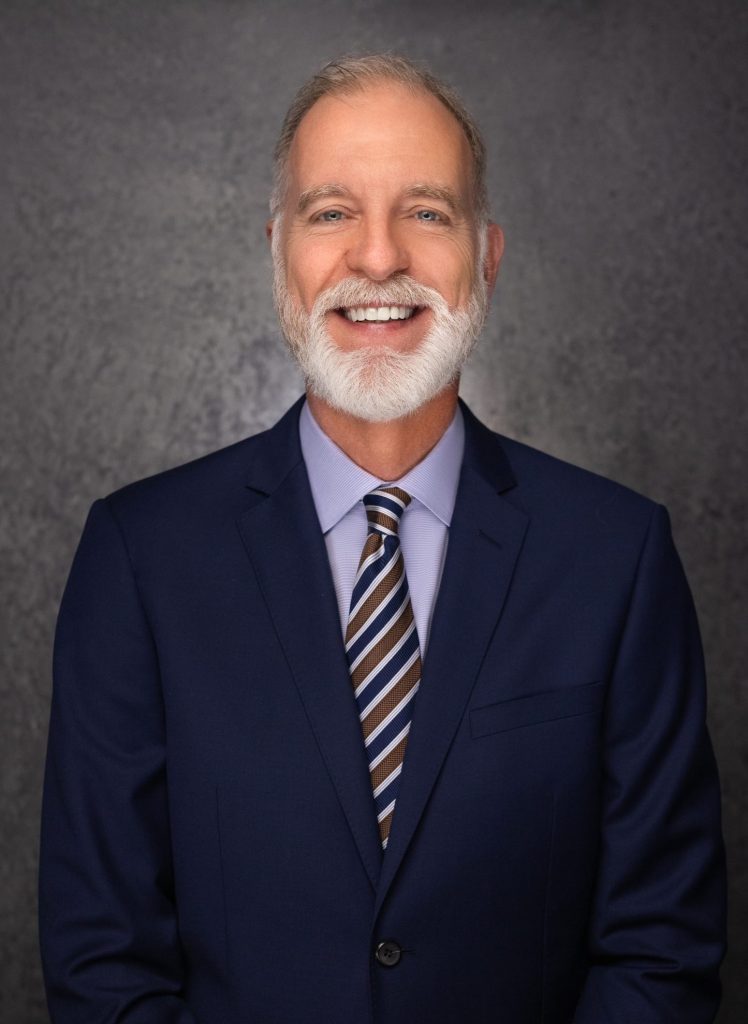A smiling older man with a white beard and mustache wears a navy blue suit, light blue shirt, and striped tie, standing in front of a dark gray textured background.