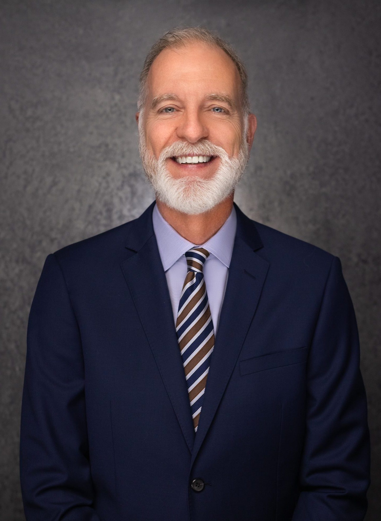 A smiling older man with a white beard and mustache wears a navy blue suit, light blue shirt, and striped tie, standing in front of a dark gray textured background.