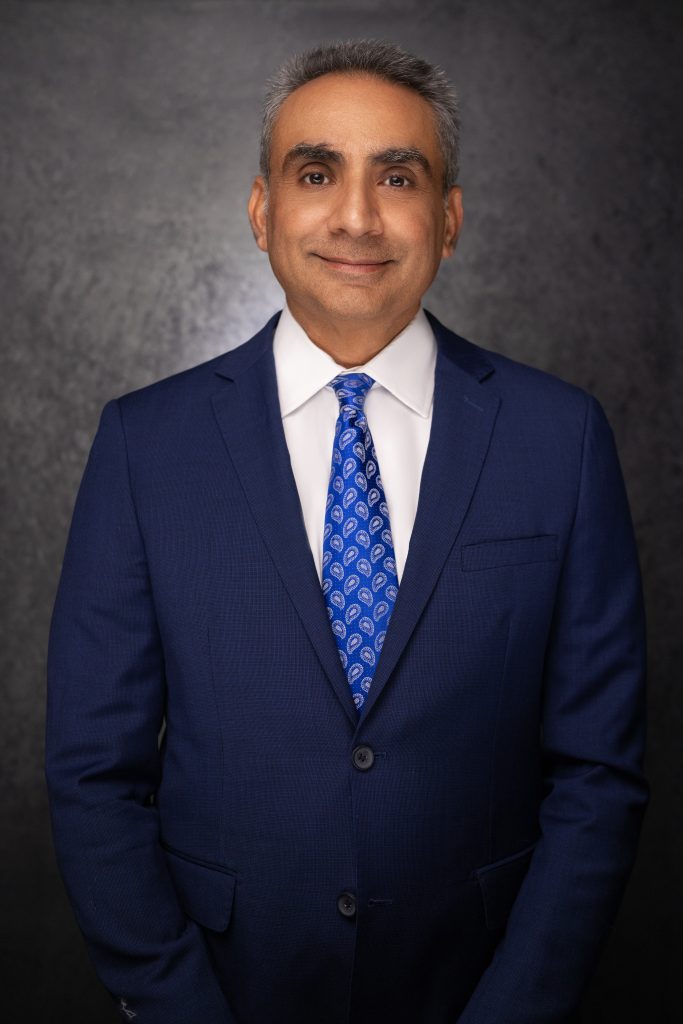 A man in a blue suit and patterned blue tie stands against a dark, textured background, smiling slightly with hands in front of him.