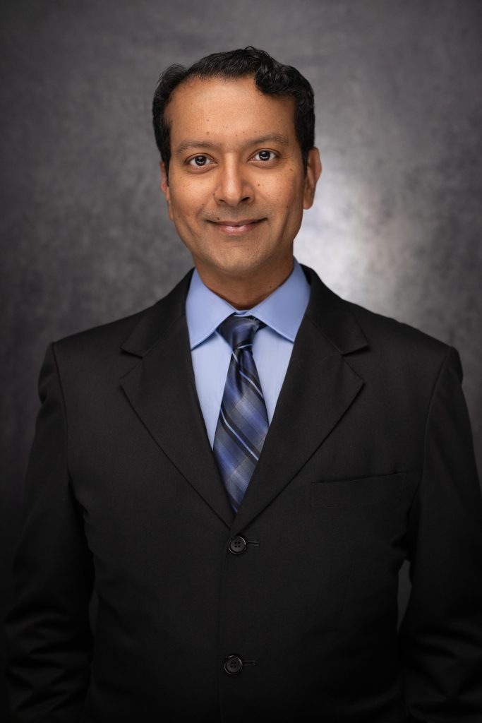 A man wearing a black suit, light blue dress shirt, and blue striped tie stands in front of a dark, textured background, smiling slightly at the camera.