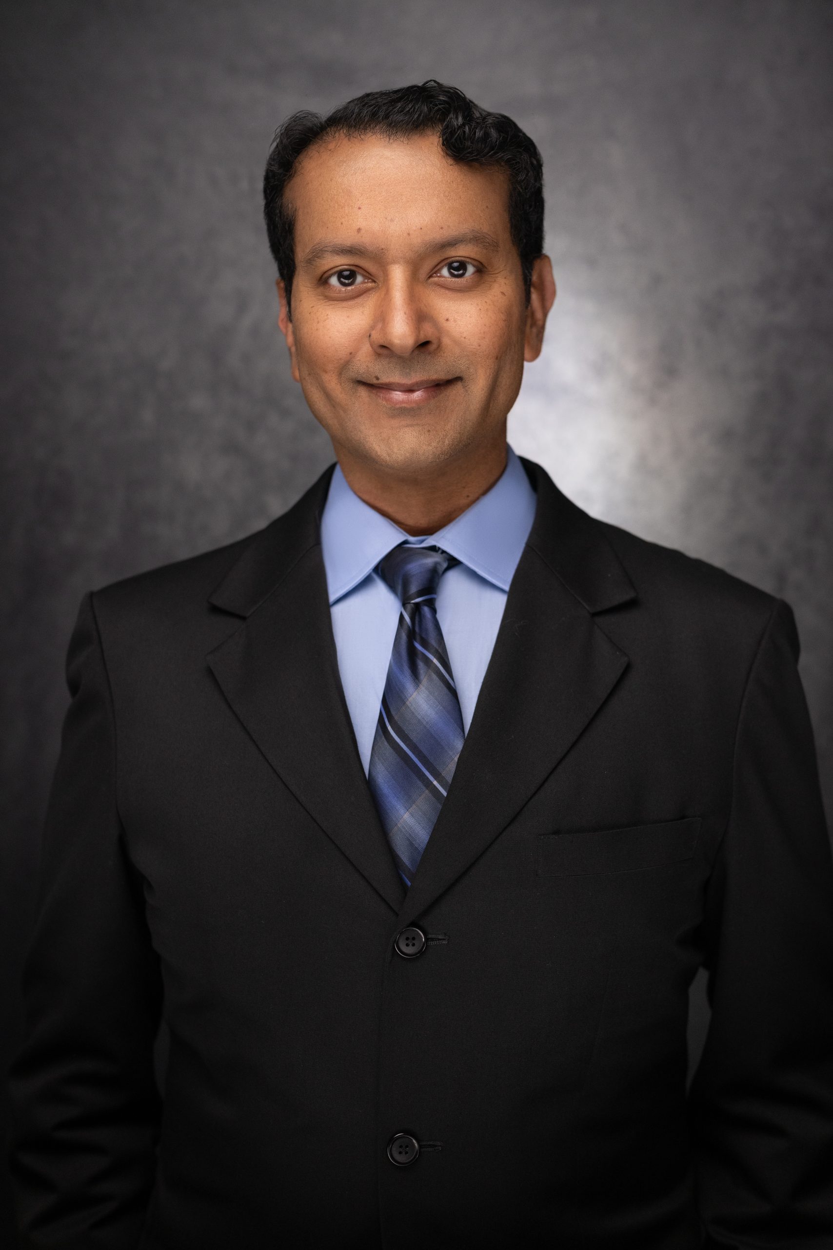 A man wearing a black suit, light blue dress shirt, and blue striped tie stands in front of a dark, textured background, smiling slightly at the camera.