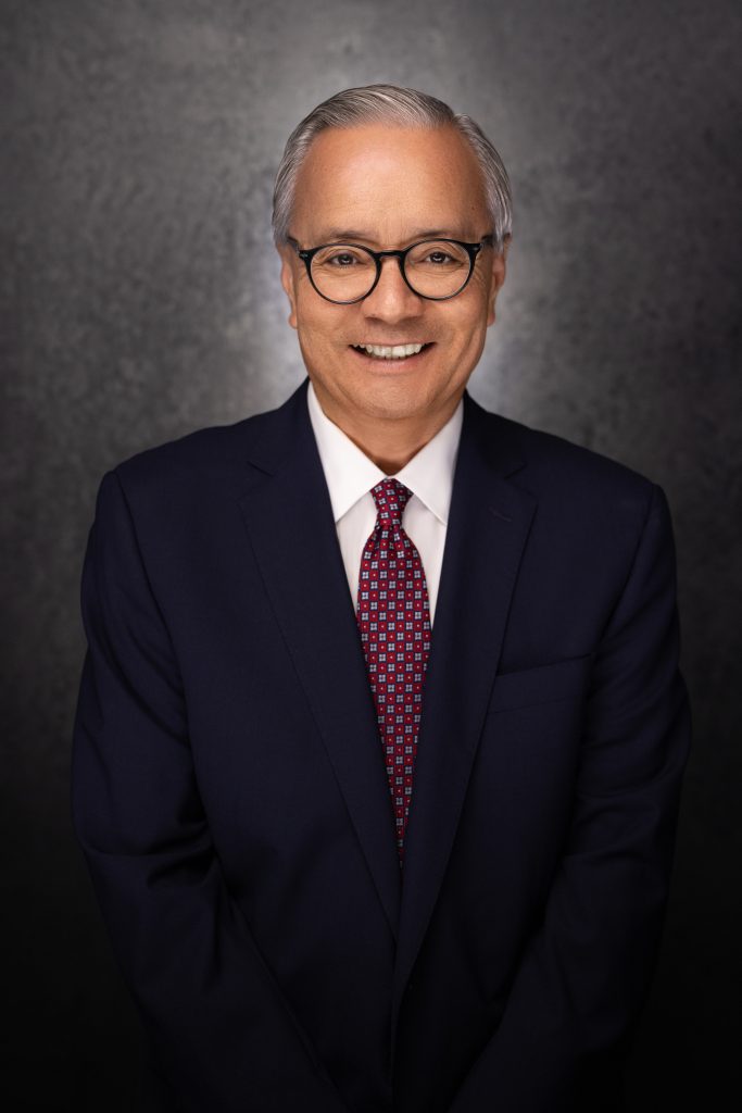 A man with short gray hair and glasses, wearing a dark navy suit, white shirt, and red patterned tie, smiles in front of a dark, textured background.
