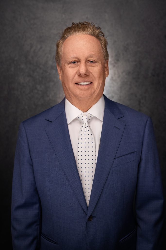 A middle-aged man with short, light hair wearing a blue suit, white dress shirt, and a white tie with black polka dots, smiling in front of a dark textured background.