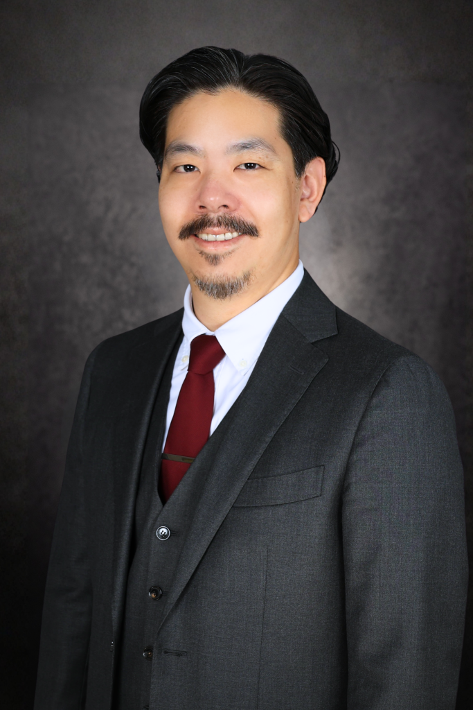 A man with dark hair and a mustache and beard, wearing a dark suit, white shirt, and a red tie, smiles at the camera against a dark, textured background.
