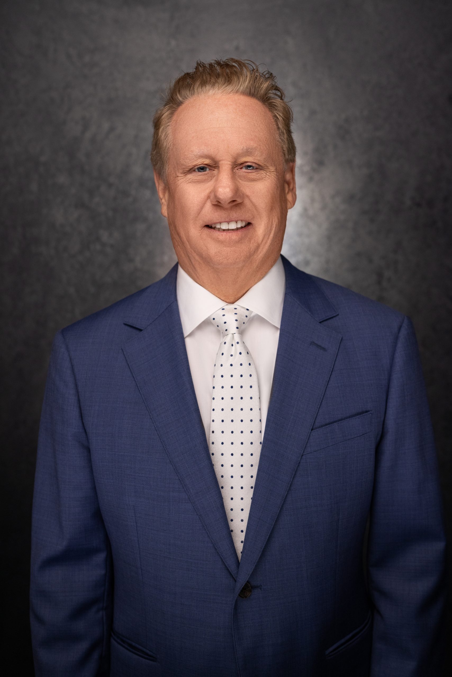 A middle-aged man with short, light hair wearing a blue suit, white dress shirt, and a white tie with black polka dots, smiling in front of a dark textured background.
