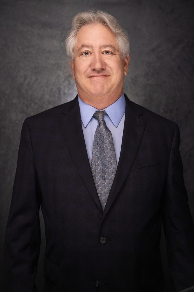 A middle-aged man with gray hair wearing a dark suit, light blue shirt, and patterned tie stands against a dark, textured background, smiling slightly.