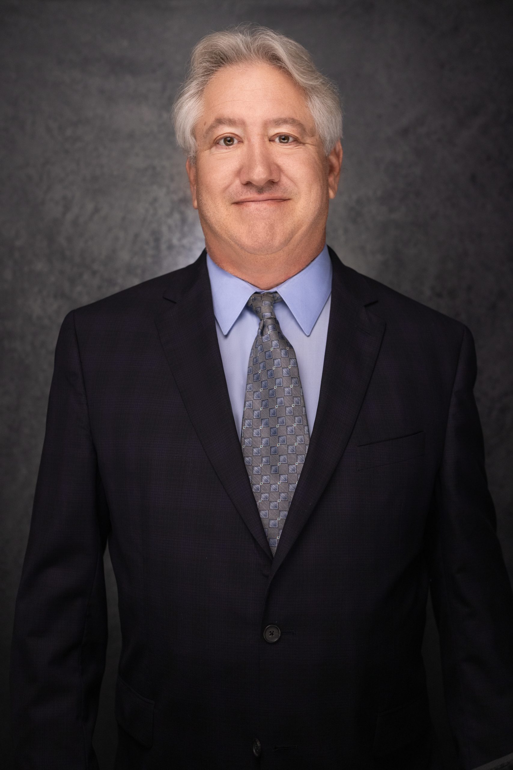 A middle-aged man with gray hair wearing a dark suit, light blue shirt, and patterned tie stands against a dark, textured background, smiling slightly.