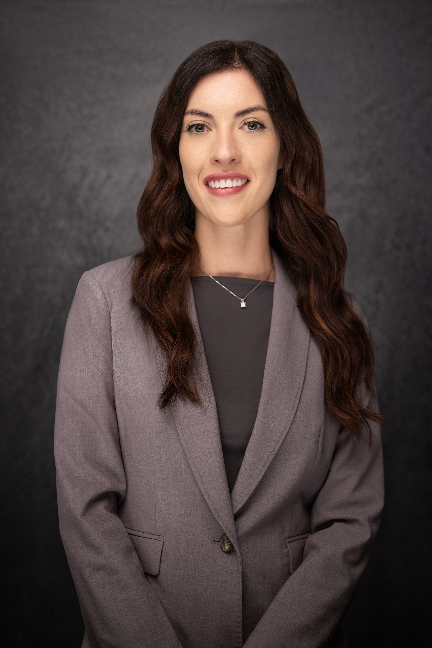A woman with long brown wavy hair, wearing a gray blazer over a dark top and a necklace, stands smiling in front of a dark textured background.