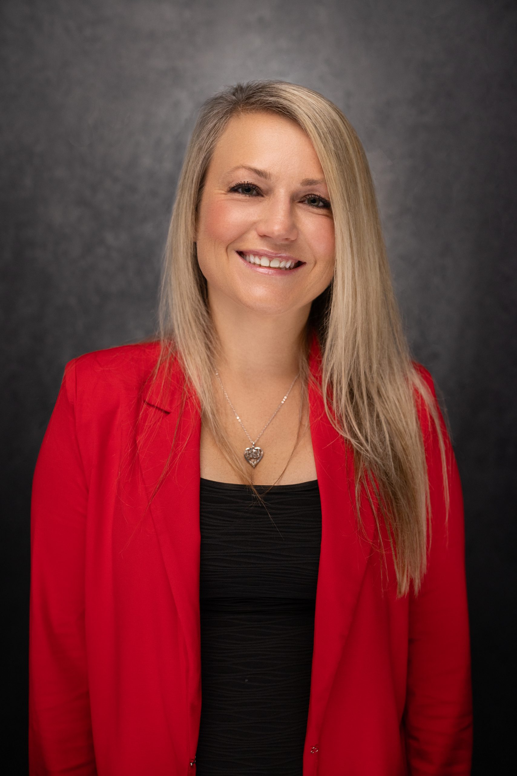 A smiling woman with long blonde hair wears a red blazer over a black top, standing in front of a dark textured background. She has a heart-shaped necklace and looks directly at the camera.