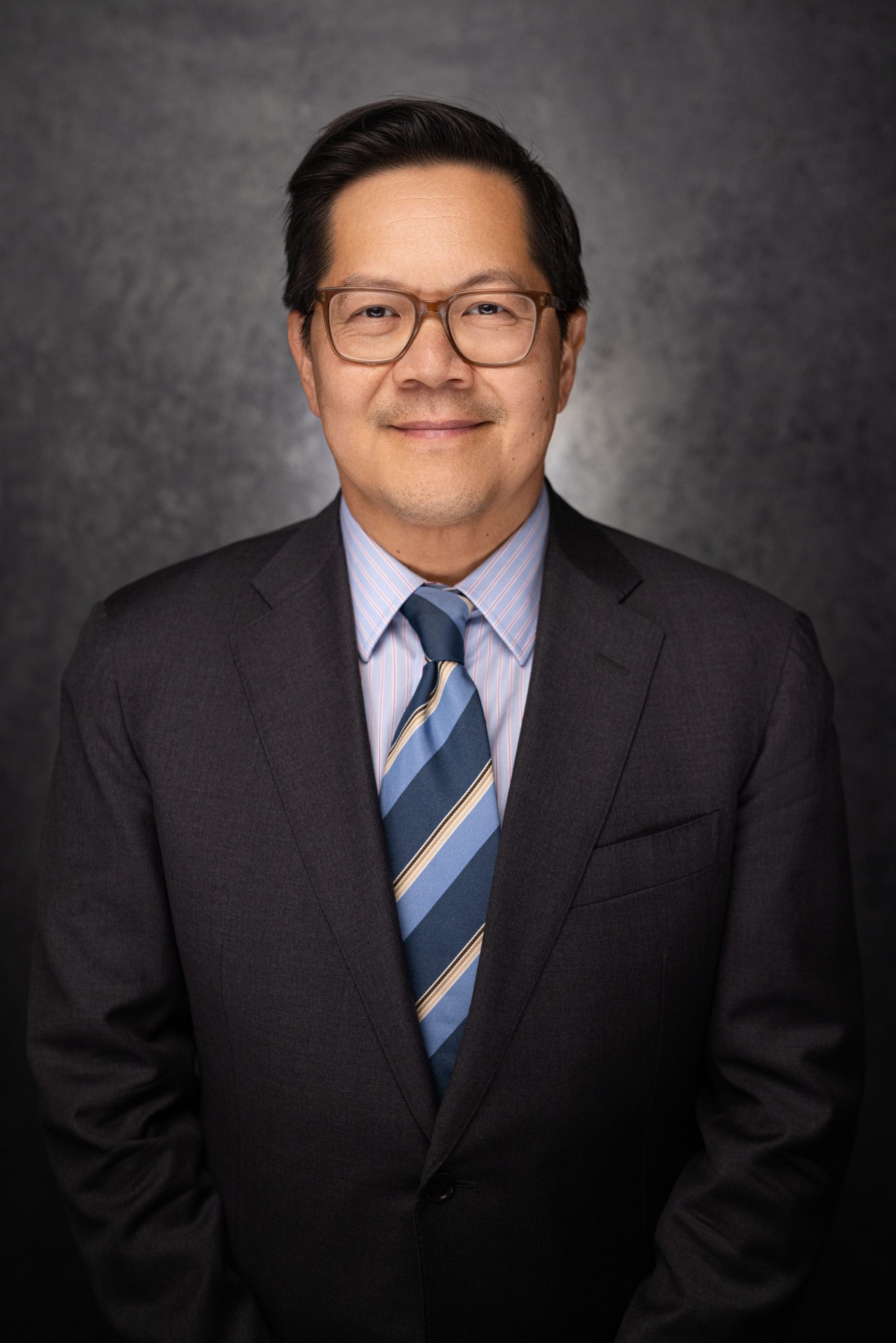 A man with short dark hair and glasses is wearing a dark suit, blue striped tie, and light blue shirt, posing in front of a dark, textured background and smiling slightly.