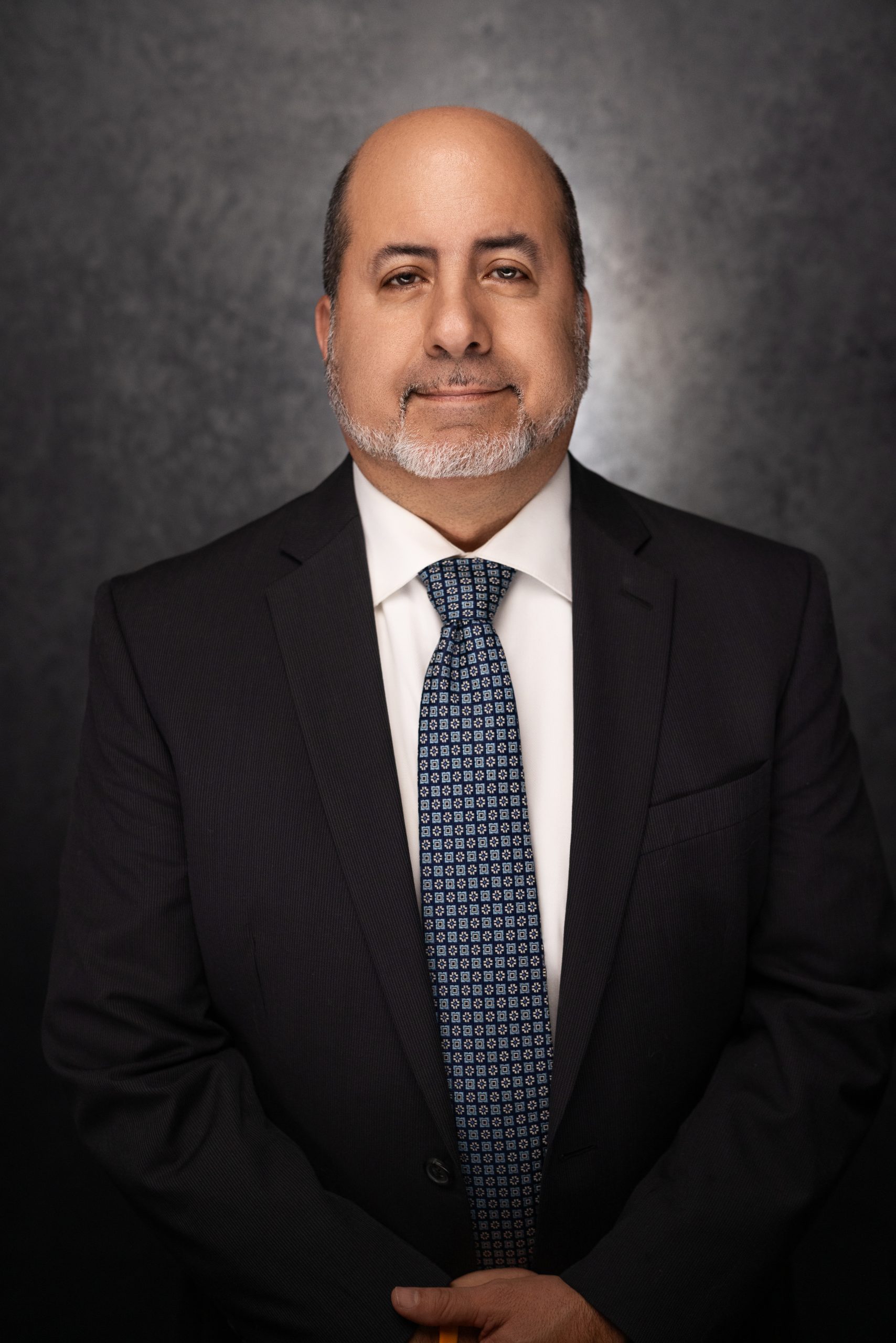 A middle-aged man with a bald head and gray beard is wearing a black suit, white shirt, and patterned blue tie, standing in front of a dark textured background. He is facing the camera with a slight smile.