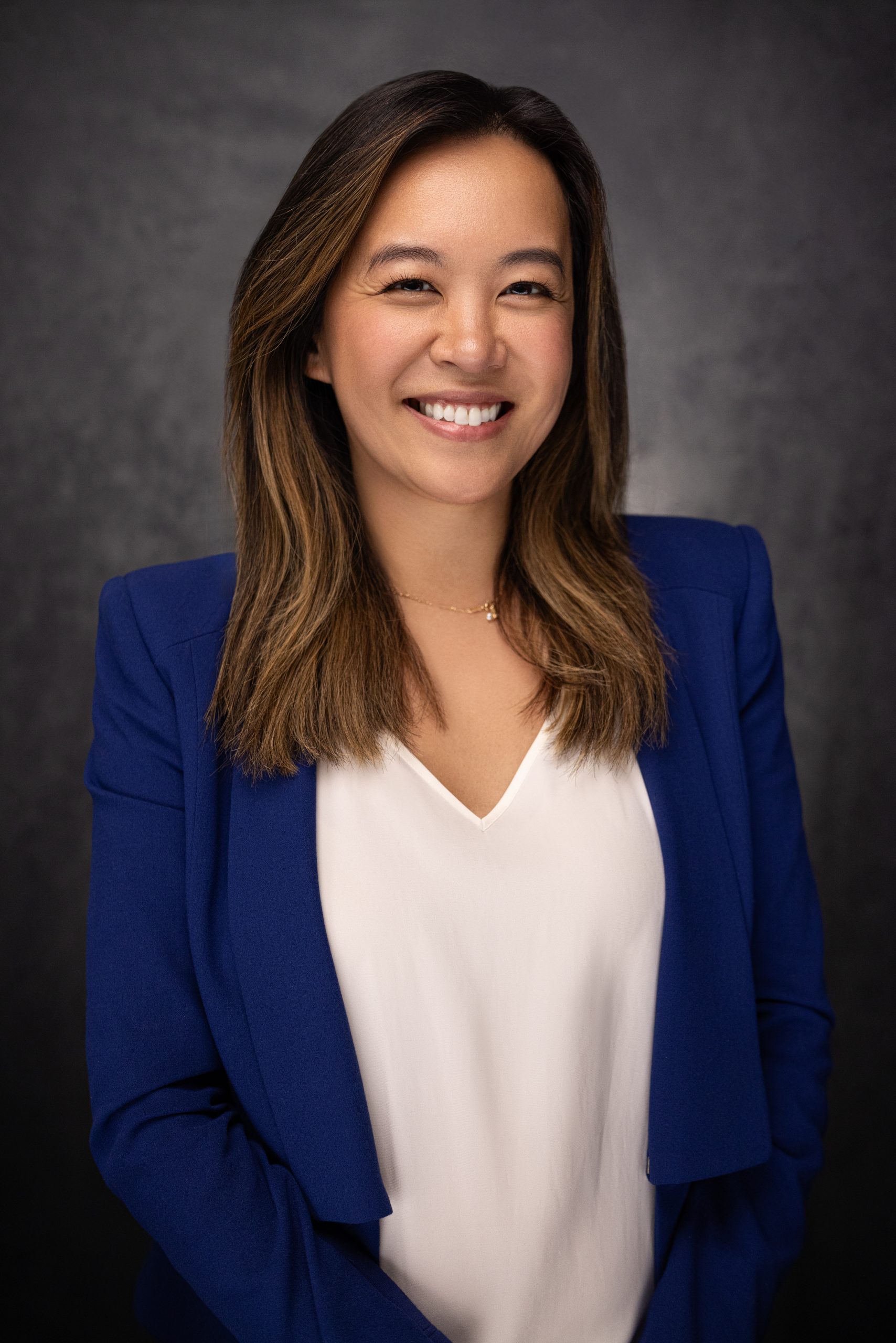 A woman with long brown hair smiles, wearing a blue blazer over a white blouse, standing against a dark, softly textured background.