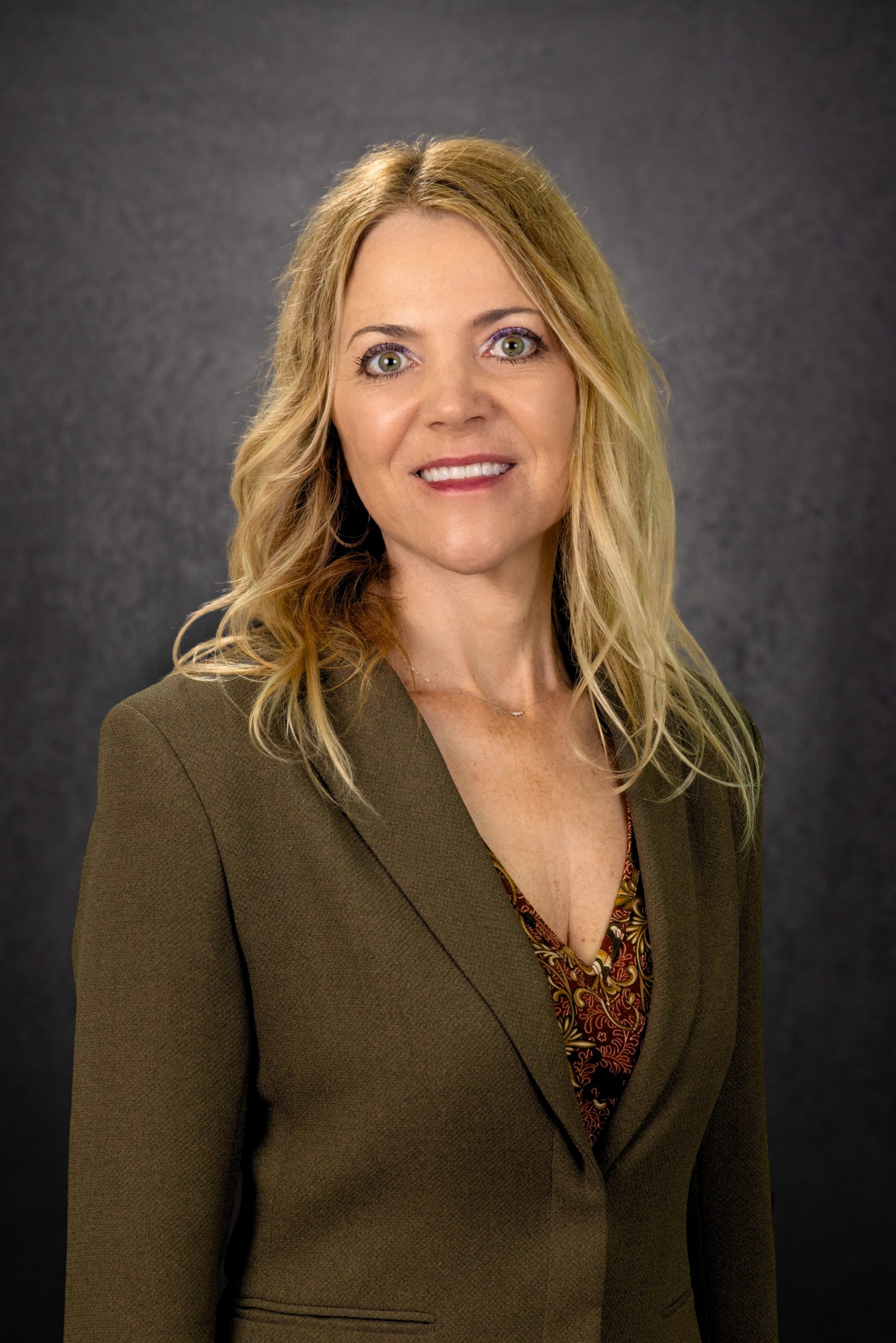 A woman with long, wavy blonde hair wearing a brown blazer and patterned top stands smiling in front of a dark gray background.