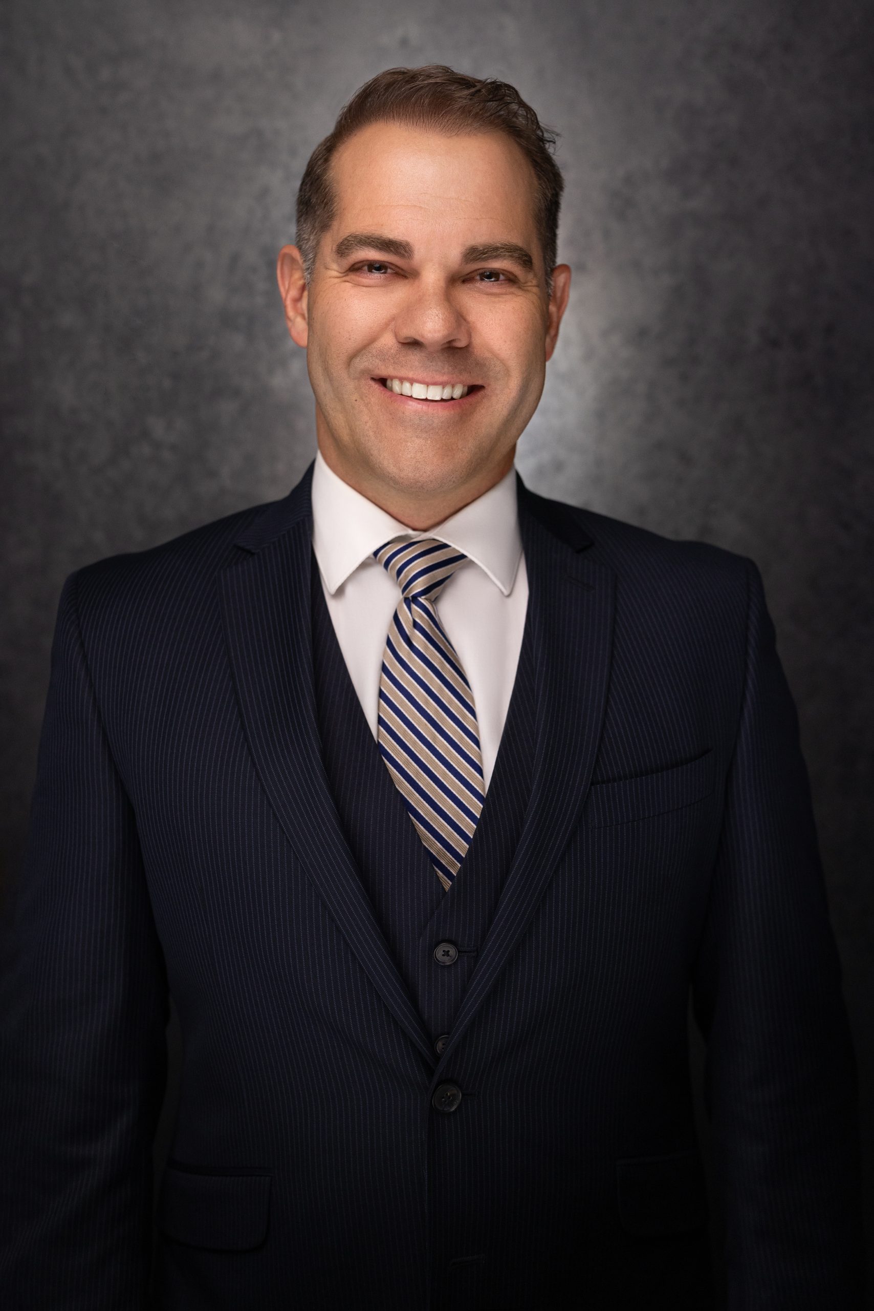 A man in a dark pinstripe suit, white shirt, and striped tie smiles at the camera against a dark, textured background.
