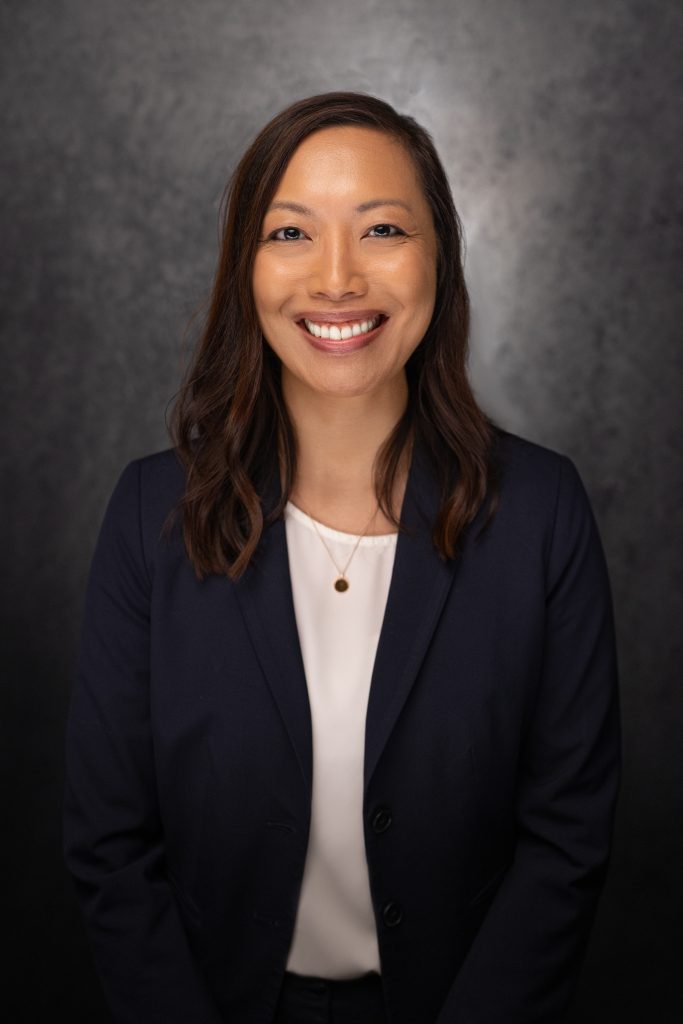 A woman with long brown hair, wearing a navy blazer over a white top and a pendant necklace, smiles at the camera against a dark, textured background.