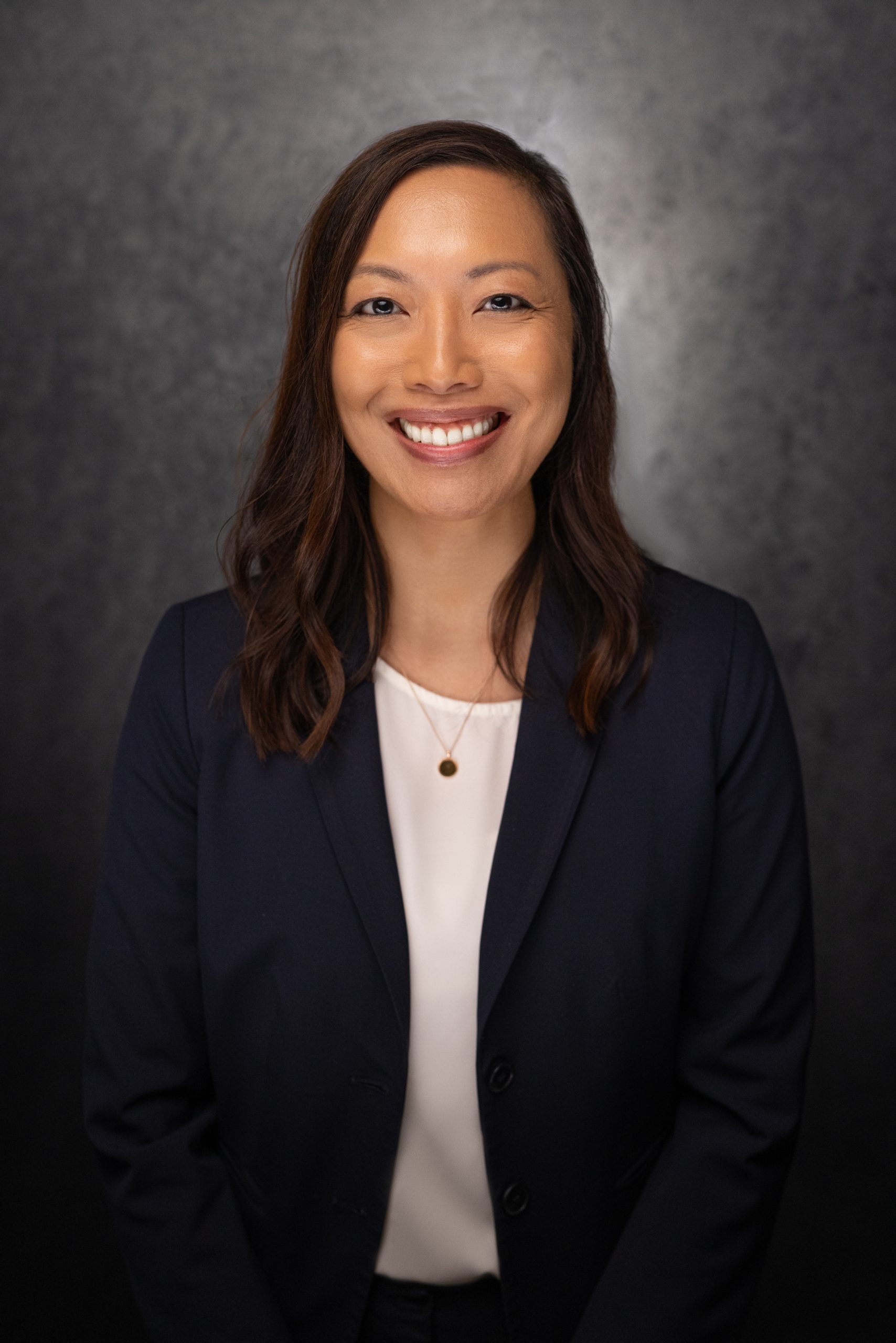 A woman with long brown hair, wearing a navy blazer over a white top and a pendant necklace, smiles at the camera against a dark, textured background.