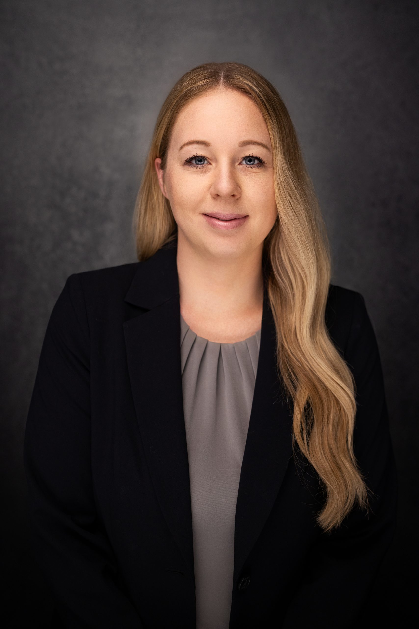 A woman with long blonde hair, wearing a black blazer and a gray blouse, poses for a professional headshot against a dark, textured background. She is smiling softly and looking at the camera.