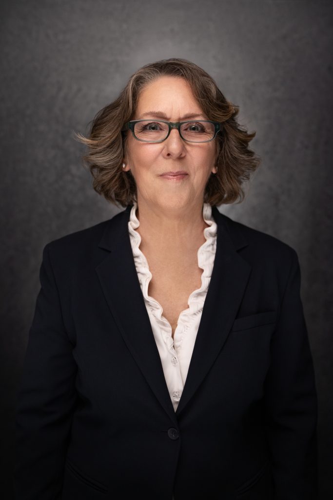 A woman with shoulder-length brown hair and glasses wears a dark blazer over a white blouse with ruffled edges, standing in front of a dark, textured background, and smiling slightly.