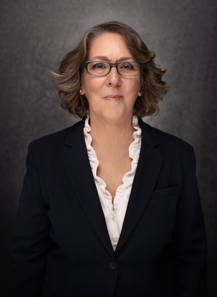 A woman with shoulder-length brown hair and glasses wears a dark blazer over a white blouse with ruffled edges, standing in front of a dark, textured background, and smiling slightly.