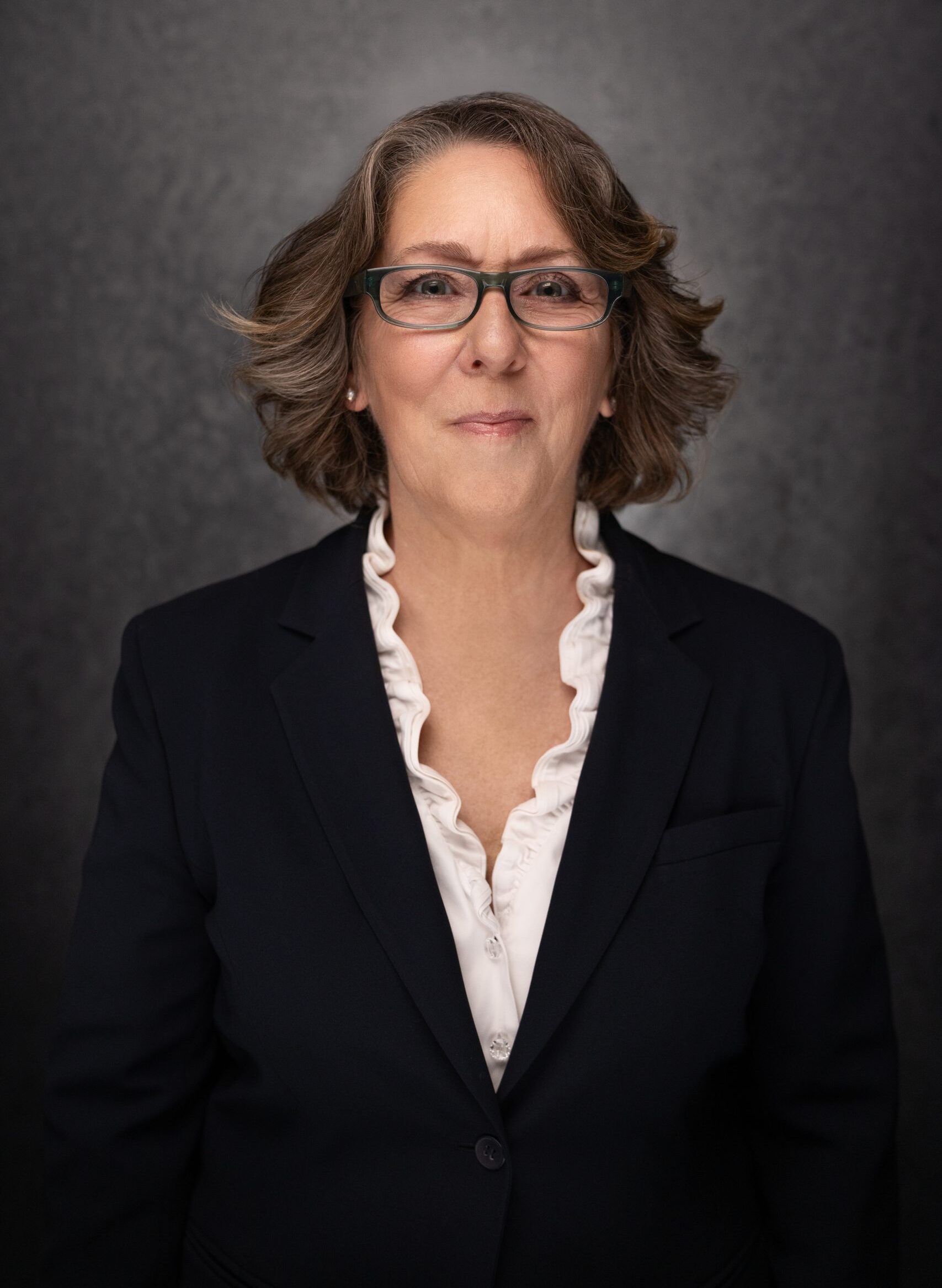 A woman with shoulder-length brown hair and glasses wears a dark blazer over a white blouse with ruffled edges, standing in front of a dark, textured background, and smiling slightly.