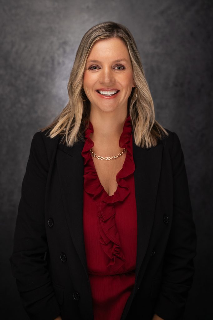 A woman with blonde hair in loose waves, wearing a black blazer over a red ruffled blouse and a gold chain necklace, smiles in front of a dark, textured background.