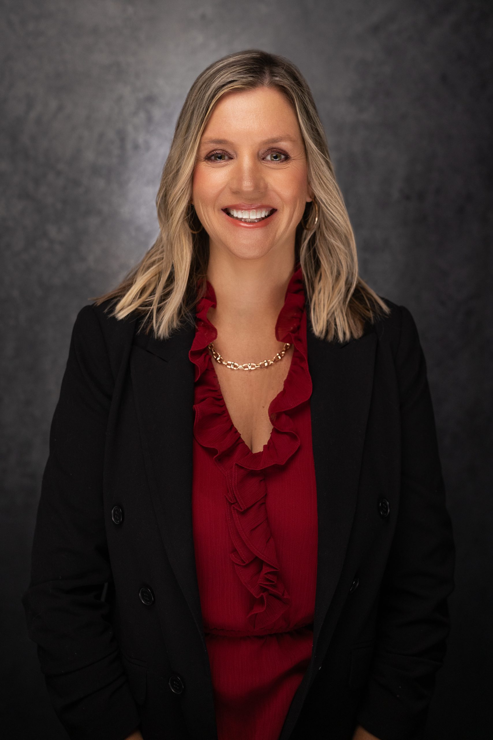 A woman with blonde hair in loose waves, wearing a black blazer over a red ruffled blouse and a gold chain necklace, smiles in front of a dark, textured background.