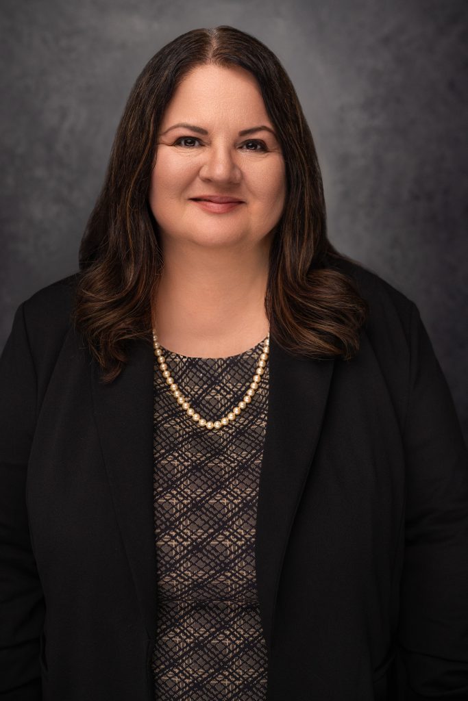 A woman with long dark hair wearing a black blazer, patterned top, and a double-strand pearl necklace smiles at the camera against a dark, textured background.