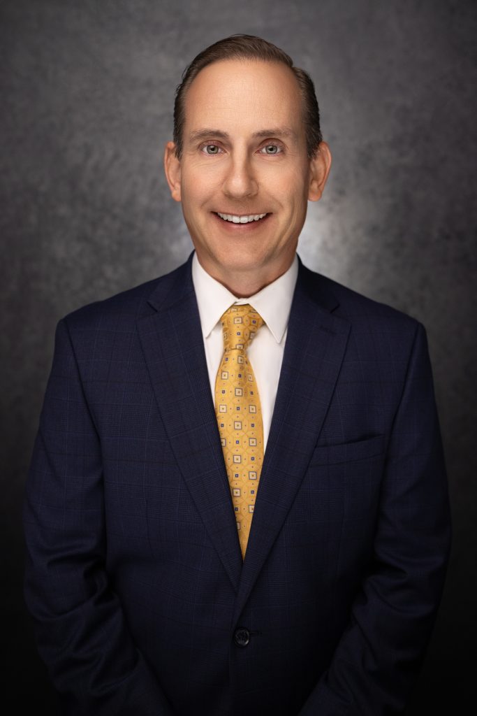 A man in a navy blue suit with a white shirt and yellow patterned tie smiles at the camera against a dark, textured background.