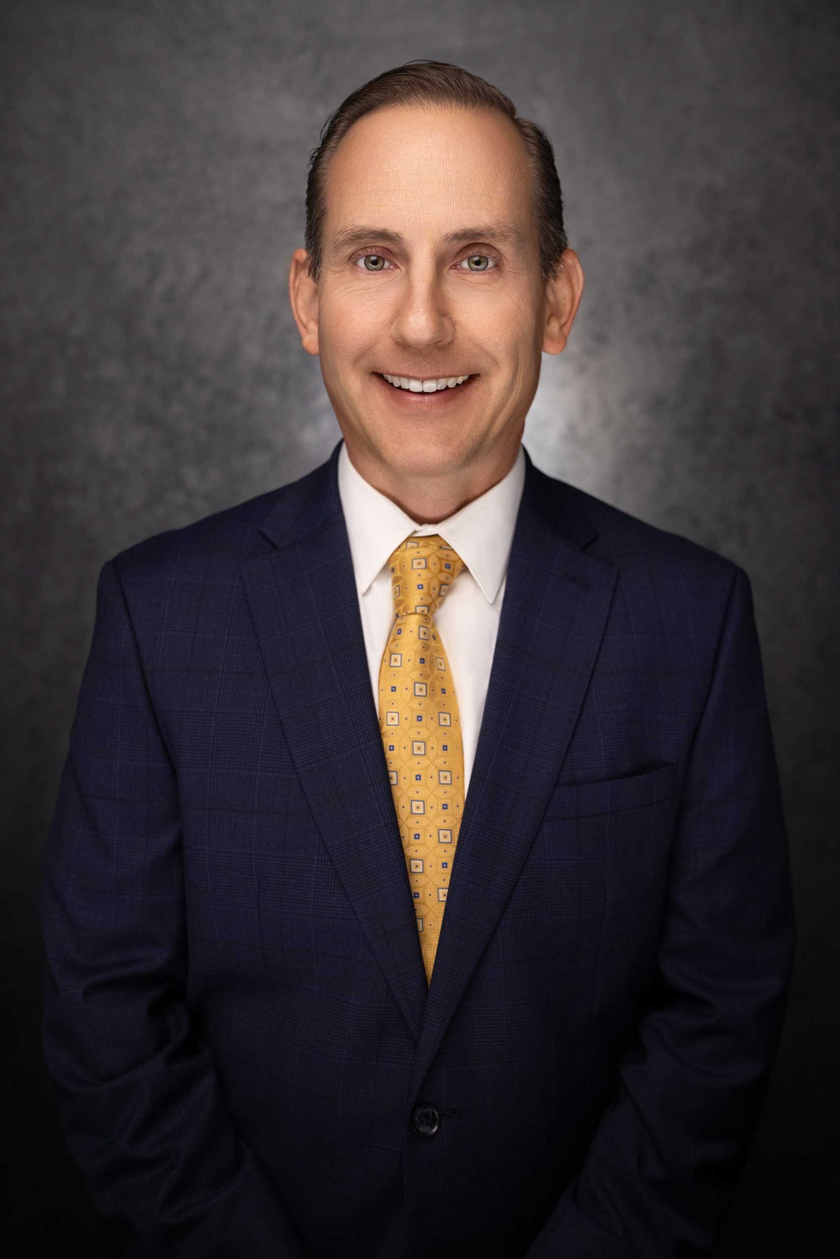 A man in a navy blue suit with a white shirt and yellow patterned tie smiles at the camera against a dark, textured background.