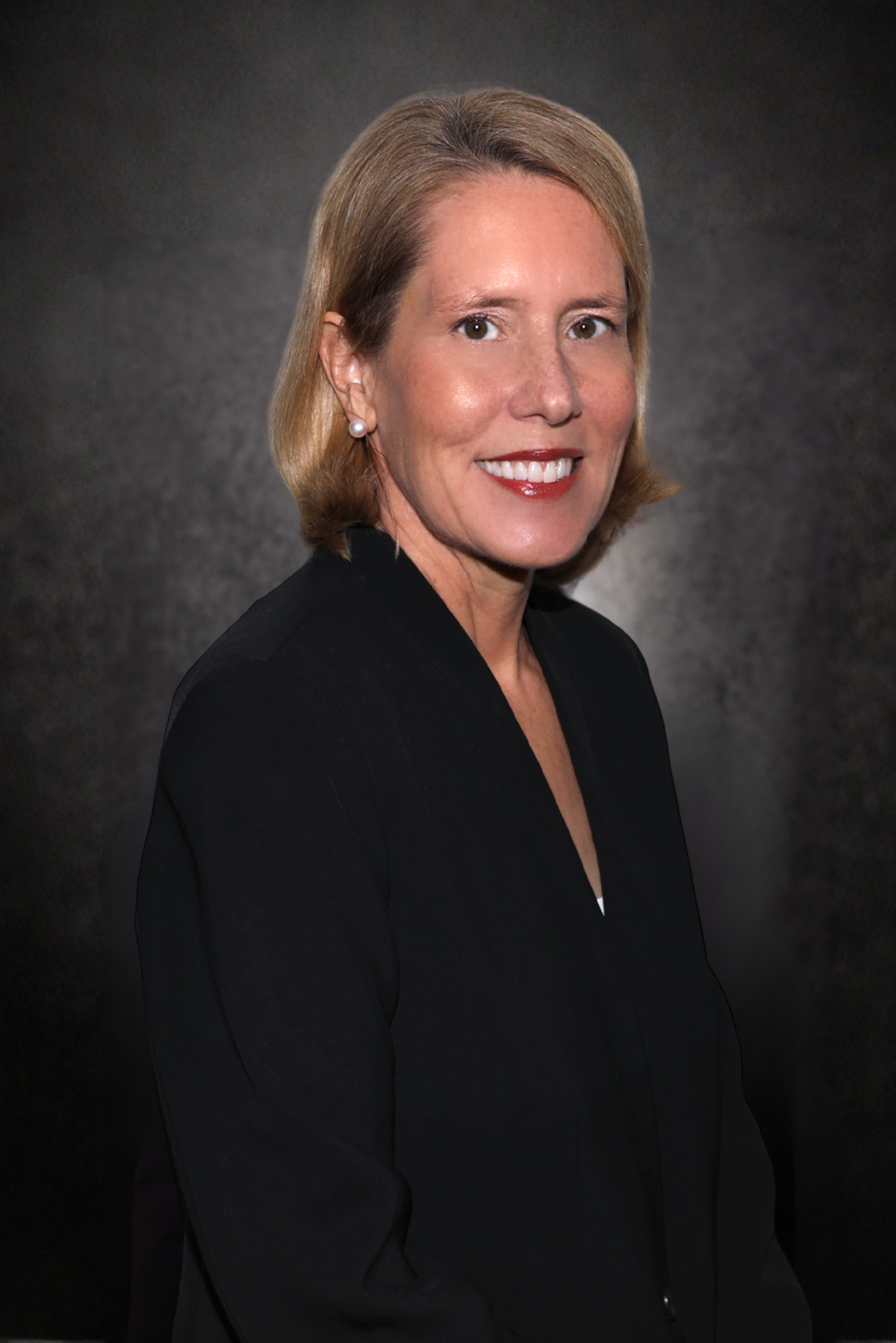A woman with short blonde hair, wearing a black blazer and small earrings, smiles at the camera against a dark, textured background.