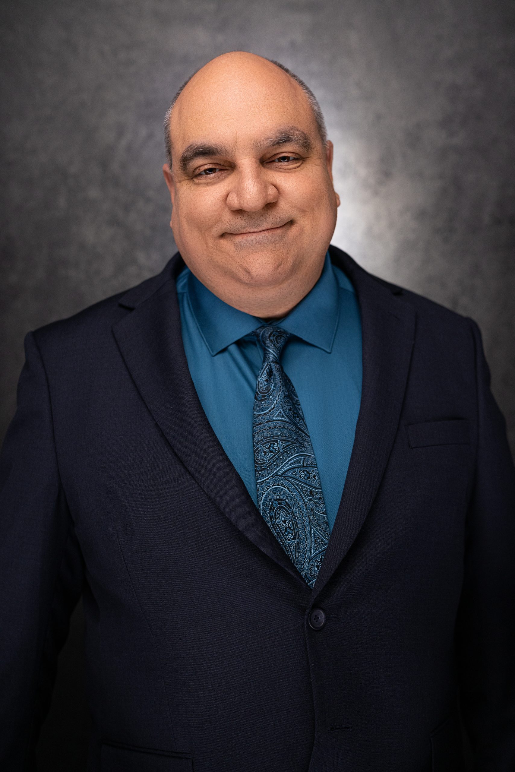 A middle-aged man with short hair smiles while wearing a dark suit, a teal dress shirt, and a patterned teal tie, posing in front of a dark, textured background.