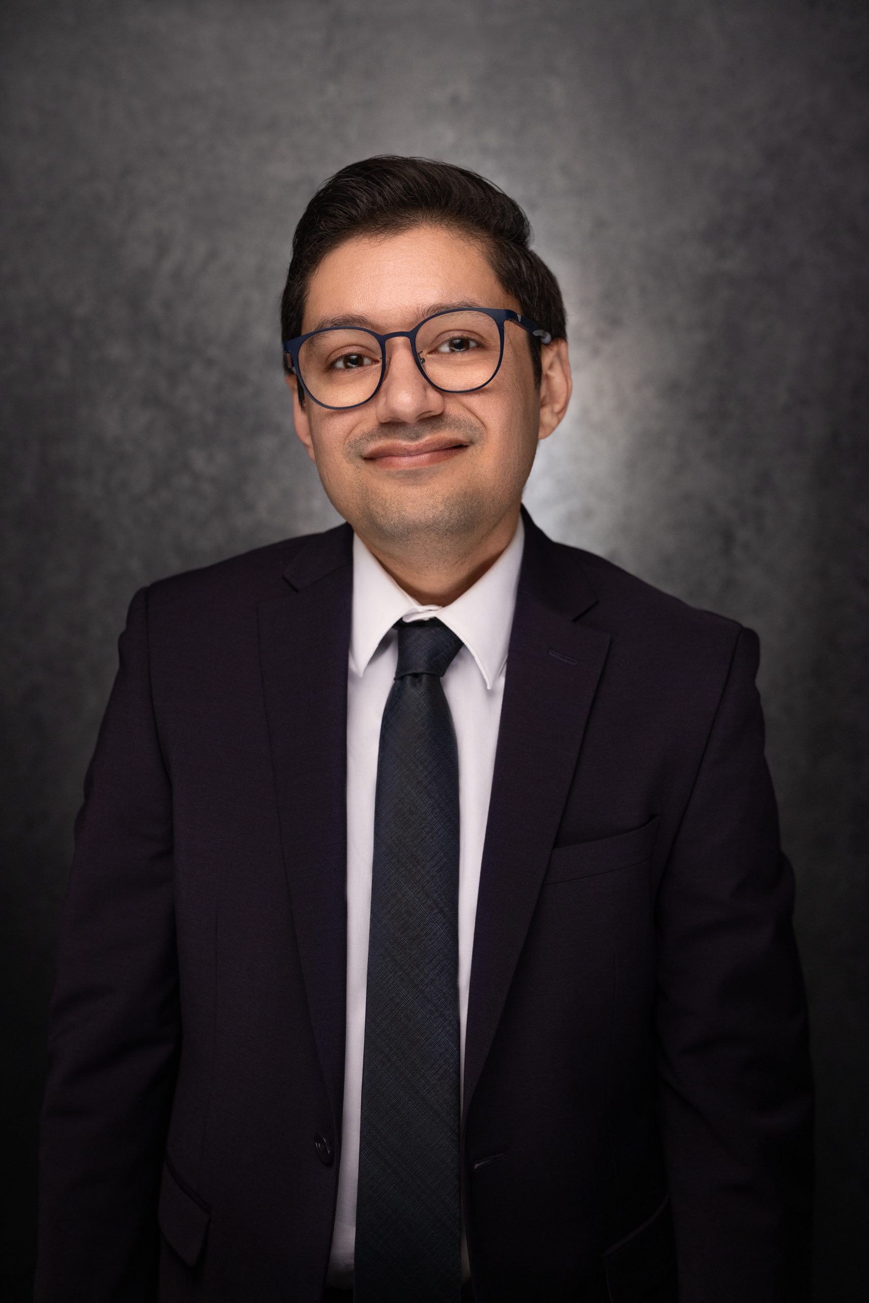 A person wearing glasses, a dark suit, white shirt, and dark tie is smiling slightly while standing in front of a dark, textured background.