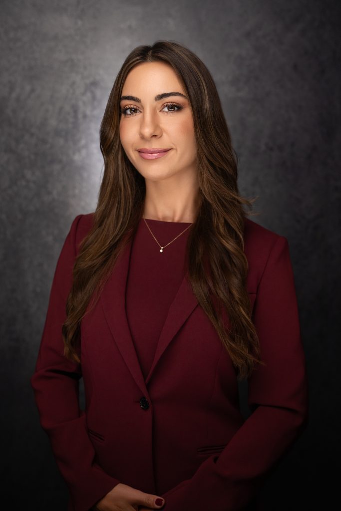 A woman with long brown hair wearing a burgundy blazer and necklace stands in front of a dark, textured background, smiling slightly with her hands clasped in front of her.